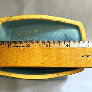 May include: A rectangular, light blue ceramic dish with a yellow rim. The dish is approximately 13 cm long. The interior has a painted design and the word "Rose" is visible. A wooden ruler is placed across the dish.