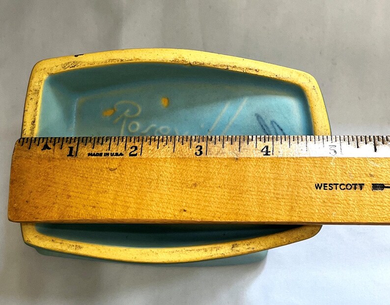 May include: A rectangular, light blue ceramic dish with a yellow rim. The dish is approximately 13 cm long. The interior has a painted design and the word "Rose" is visible. A wooden ruler is placed across the dish.