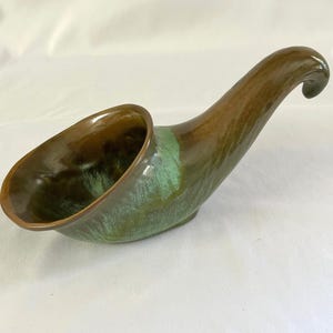 Pode incluir: Um prato de servir em cerâmica com formato de chifre. Apresenta um esmalte marrom e verde, com uma alça curva e uma tigela larga e aberta. A cor transita do marrom para o verde, criando uma aparência única e artística. Provavelmente usado para servir alimentos.