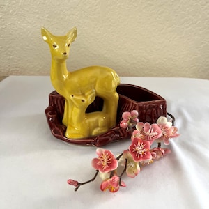 May include: A ceramic deer figurine in yellow, featuring a mother deer and fawn, set within a burgundy planter. Pink and white artificial flowers are arranged nearby, creating a decorative display.
