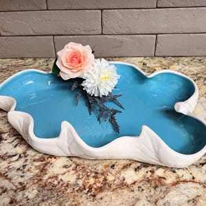 May include: A decorative, cloud-shaped ceramic dish with a glossy, light blue interior and a white, textured rim. The dish is adorned with a pink rose, a white chrysanthemum, and sprigs of blue-gray foliage. The dish is designed for decorative purposes.
