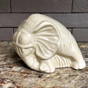 May include: Cream-colored ceramic elephant figurine. The elephant is depicted in a resting position, with its trunk curled and ears spread. The figurine has a glossy finish and detailed features, showcasing the texture of the elephant's skin.
