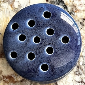 May include: A circular, dark blue ceramic flower frog featuring a glossy, speckled glaze finish. The top surface is punctuated by eleven evenly spaced, circular holes designed for arranging individual flower stems in a floral display.