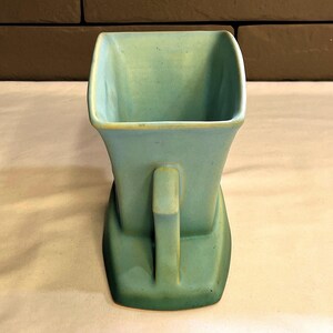 May include: A vintage, square-shaped vase in a muted teal colour with a handle. The vase has a unique, stepped base and a slightly flared top. The handle is rectangular and positioned on the side. The vase is made of ceramic.