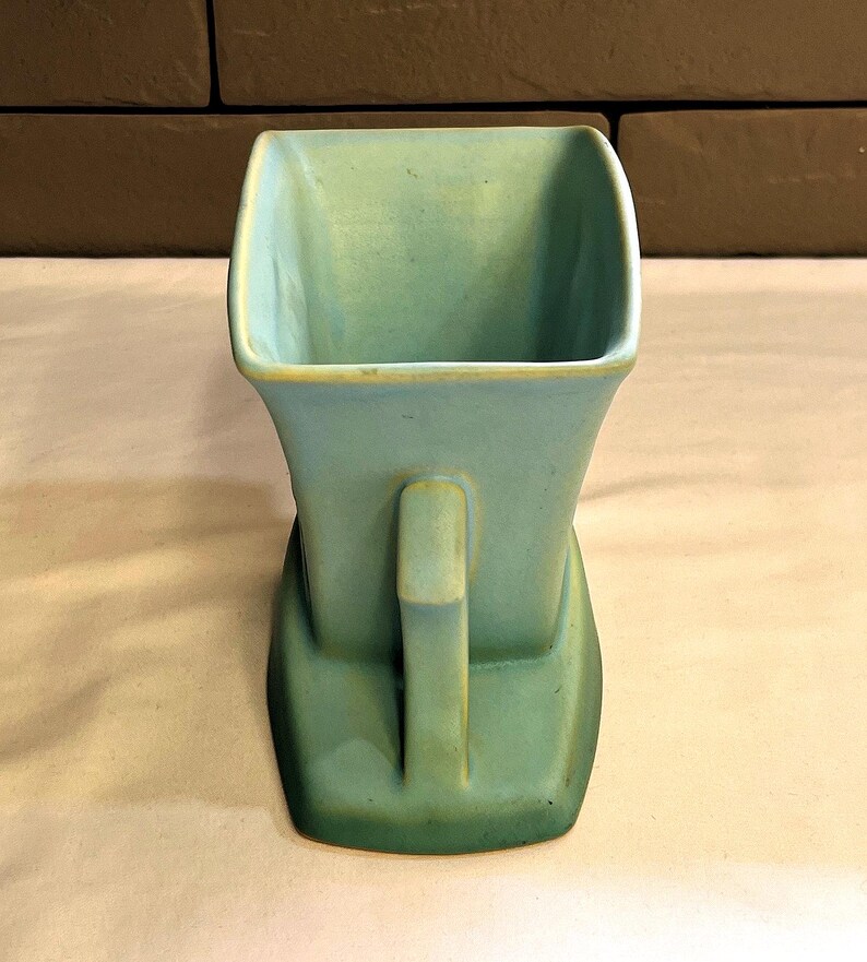 May include: A vintage, square-shaped vase in a muted teal colour with a handle. The vase has a unique, stepped base and a slightly flared top. The handle is rectangular and positioned on the side. The vase is made of ceramic.