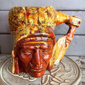 May include: A ceramic figural mug shaped like a person wearing a feathered headdress, featuring a glossy glaze in shades of deep amber, brown, and yellow. The handle is sculpted to resemble a feather, and the mug has a detailed, textured surface.