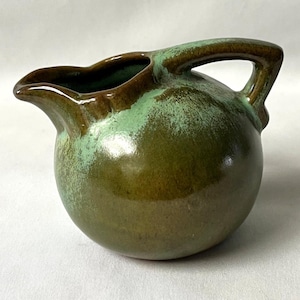 Pode incluir: Jarro de cerâmica pequeno e redondo com asa e bico. O jarro é esmaltado em tons de verde e castanho, com um efeito manchado. A asa e a borda são de um castanho mais escuro.