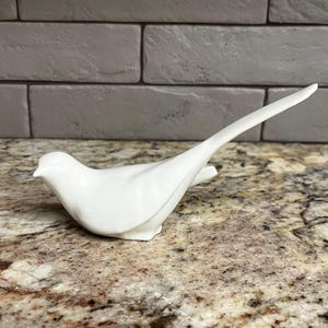 May include: A white ceramic bird figurine with a long, upward-curving tail. The bird is perched on a speckled countertop, with a gray brick wall in the background. The figurine is smooth and glossy, showcasing a minimalist design.