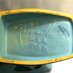 May include: Close-up of the base of a vintage ceramic vase. The vase is turquoise with a yellow rim. The text "Roseville U.S.A. 756" is visible.