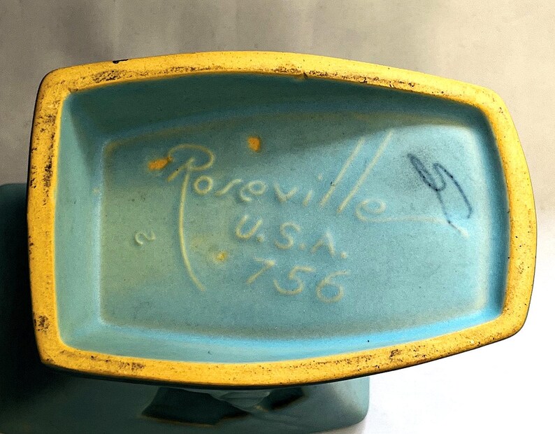 May include: Close-up of the base of a vintage ceramic vase. The vase is turquoise with a yellow rim. The text "Roseville U.S.A. 756" is visible.