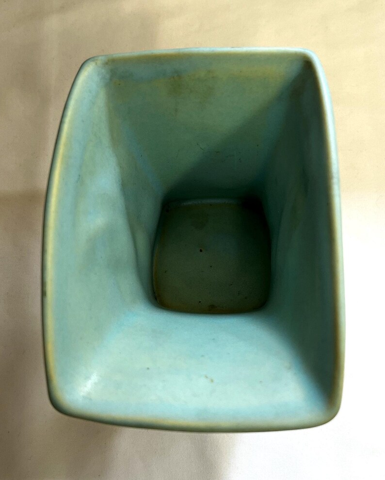 May include: A square turquoise ceramic vase with rounded corners. The interior is a darker shade of turquoise. The vase has a smooth, glossy finish and appears handmade. The design is simple and modern. The vase is approximately 15 cm tall.