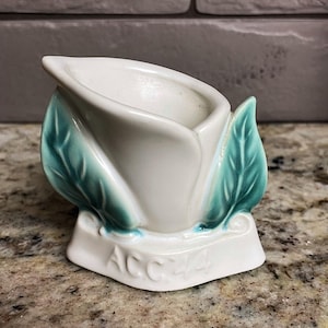 Peut inclure: Un vase à bourgeon en céramique blanche en forme de fleur, avec des feuilles et des accents vert turquoise. La base porte le texte "ACC 44". Le vase est sur une surface mouchetée.