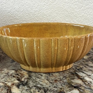 May include: A ceramic bowl with a speckled, golden-yellow glaze. The bowl has a ribbed exterior and a rounded shape, suitable for serving or display. The bowl sits on a small, circular base.