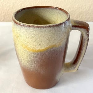 May include: A ceramic mug with a rectangular handle. The mug has a gradient design, transitioning from a dark brown at the base to a beige color at the rim. A thin yellow band separates the two colors. The mug is suitable for hot or cold beverages.