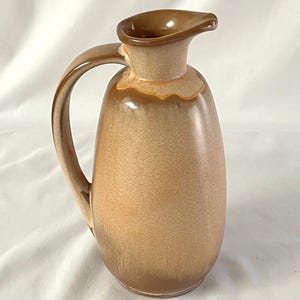 Pode incluir: Um jarro de cerâmica com uma alça e um bico. O jarro tem um corpo arredondado e uma cobertura marrom clara que escurece em direção à base. A alça e o bico são de um tom mais escuro.