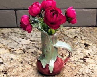 May include: A small, handcrafted ceramic vase with a handle, glazed in green and red hues, holds a bouquet of vibrant pink peonies. The vase has a unique, organic shape, adding a touch of artistic flair to the floral arrangement.