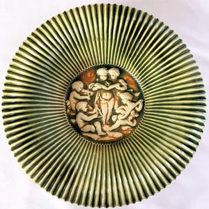 Könnte beinhalten: Ein dekorativer grüner Keramikteller mit geripptem, sonnenförmigem Design. Die Mitte zeigt eine Reliefskulptur von Cherubinen in Creme- und Brauntönen. Der Teller ist rund und hat einen Durchmesser von ca. 25 cm.