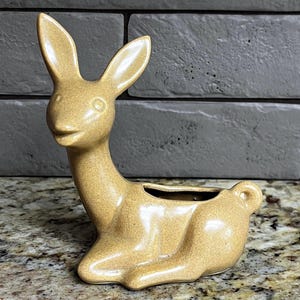 May include: A tan ceramic bunny planter with a small opening for plants. The bunny is in a seated position with its ears upright. The planter has a glossy finish and is on a speckled surface.