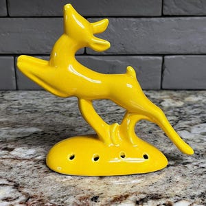 May include: A bright yellow ceramic figurine of a leaping deer. The deer is depicted in a dynamic pose with its head tilted upward and legs extended. The base is oval-shaped and features four small, evenly spaced circular holes along the front side.