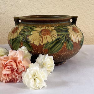 Peut inclure: Un vase en céramique marron avec un motif floral représentant des fleurs jaunes et blanches et des feuilles vertes. Le vase a deux petites poignées et est décoré d'un motif texturé. Le vase mesure environ 20 cm de diamètre.