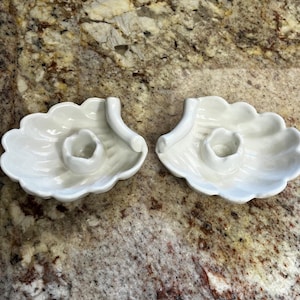 Peut inclure: Deux bougeoirs en céramique blanche. Chaque support a la forme d'une feuille stylisée avec une coupelle centrale pour une bougie et une poignée incurvée. Les supports sont sur une surface marron mouchetée.