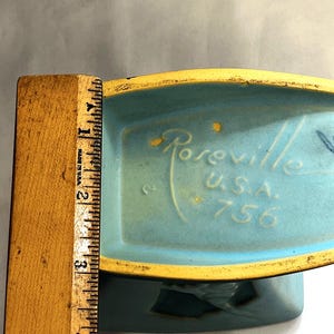 May include: A vintage turquoise ceramic planter with a yellow rim. The base is marked "Roseville U.S.A. 756". A wooden ruler shows the planter is approximately 7.6 cm tall.