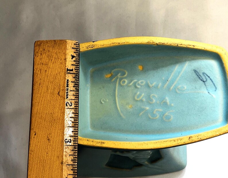 May include: A vintage turquoise ceramic planter with a yellow rim. The base is marked "Roseville U.S.A. 756". A wooden ruler shows the planter is approximately 7.6 cm tall.