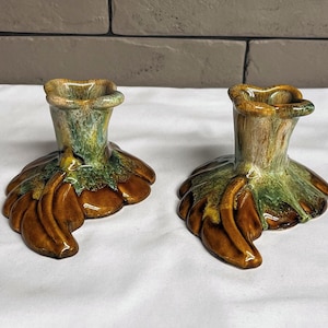 May include: Two ceramic candle holders with a unique design. The holders have a brown, green, and tan color scheme. The base of each holder is shaped like a stylized leaf, and the top has a small opening for a candle. The candle holders are on a white surface.