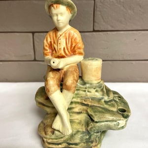 May include: A ceramic figurine of a young person wearing an orange shirt and shorts, seated on a textured, green and brown base. The figure is wearing a green hat and holding a small object. The figurine has a vintage aesthetic.