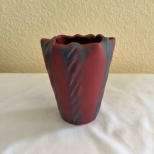 May include: A maroon-colored vase with a textured, wavy design. The vase has a flared top and a cylindrical body. Dark blue accents highlight the textured pattern. The vase is sitting on a white surface.