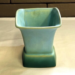 May include: A square-shaped, vintage vase in shades of blue and green. The vase has a flared top and a rectangular base. The colours transition from a light blue at the top to a darker green at the base, with a gold accent.