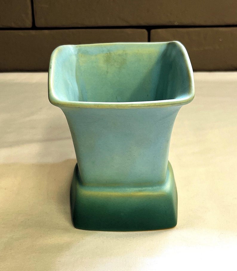 May include: A square-shaped, vintage vase in shades of blue and green. The vase has a flared top and a rectangular base. The colours transition from a light blue at the top to a darker green at the base, with a gold accent.