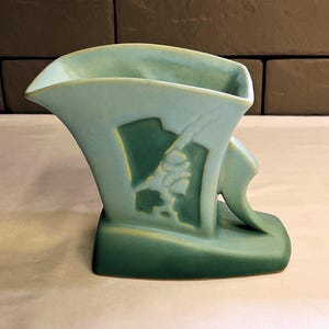 May include: A vintage art deco vase in shades of green. The vase features a stylized design with a curved handle and a decorative relief. The vase has a wide opening and a rectangular base. The overall design is symmetrical and elegant.