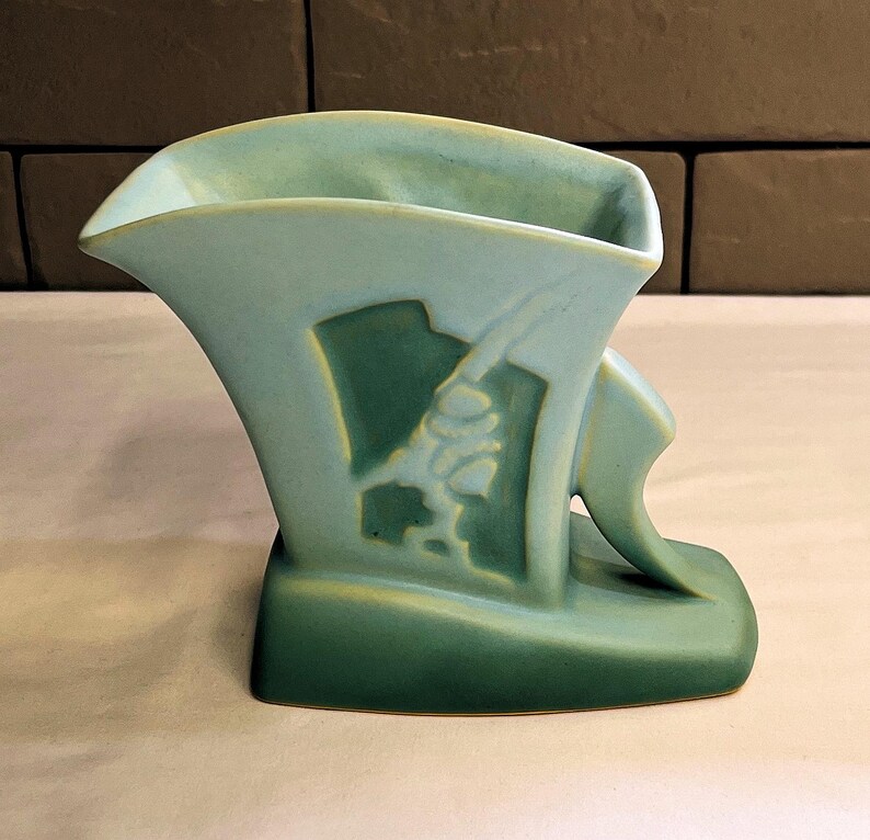 May include: A vintage art deco vase in shades of green. The vase features a stylized design with a curved handle and a decorative relief. The vase has a wide opening and a rectangular base. The overall design is symmetrical and elegant.