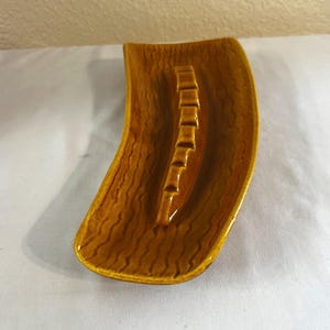 Op de afbeelding: Een vintage, gebogen asbak in een warme, aardse tint. De asbak heeft een textuur die lijkt op houtnerf en heeft een centrale groef met meerdere rustplaatsen voor sigaretten. Het algehele ontwerp is een retro-stijl.
