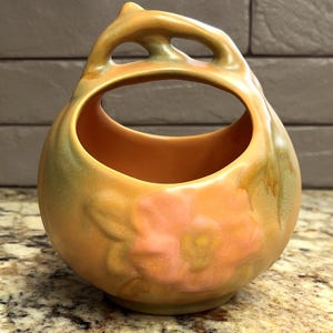 May include: A round ceramic basket with a high arched handle featuring a cutout design. The vessel is finished in a warm peach and orange glaze with subtle green shading and a raised, embossed floral motif on the front center of the body.