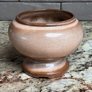 Pode incluir: Uma tigela de cerâmica com pedestal, corpo arredondado e abertura larga. A tigela é uma mistura de tons marrons e rosa, com borda e base marrons mais escuras. A superfície texturizada adiciona interesse visual.