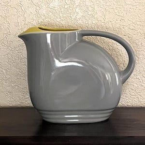May include: A glossy gray ceramic pitcher with a rounded body, a curved handle, and a distinct spout. The interior of the pitcher features a contrasting yellow glaze visible at the rim, while the exterior base is accented with three horizontal ridges.