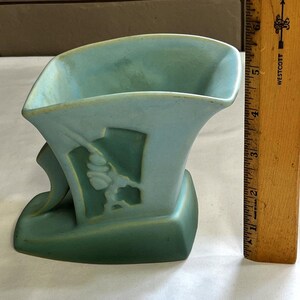 May include: A two-tone, art deco-style vase in shades of teal and green. The vase features a stylized floral design on its side. A wooden ruler is positioned next to the vase, providing a size reference.