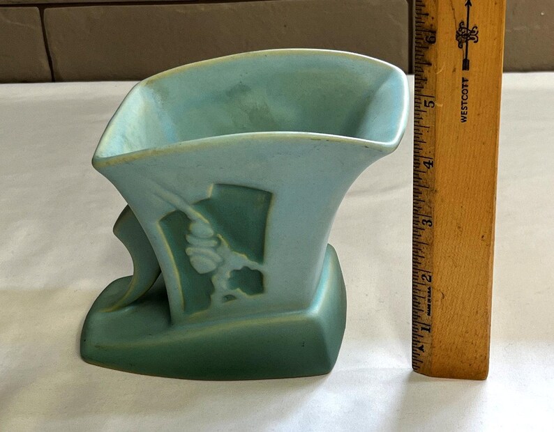 May include: A two-tone, art deco-style vase in shades of teal and green. The vase features a stylized floral design on its side. A wooden ruler is positioned next to the vase, providing a size reference.