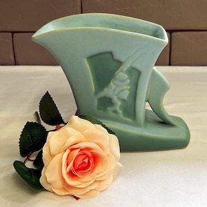 May include: A pale blue art deco vase with a relief design, next to a peach-coloured rose with green leaves. The vase has a unique shape and a matte finish, suitable for home decor.