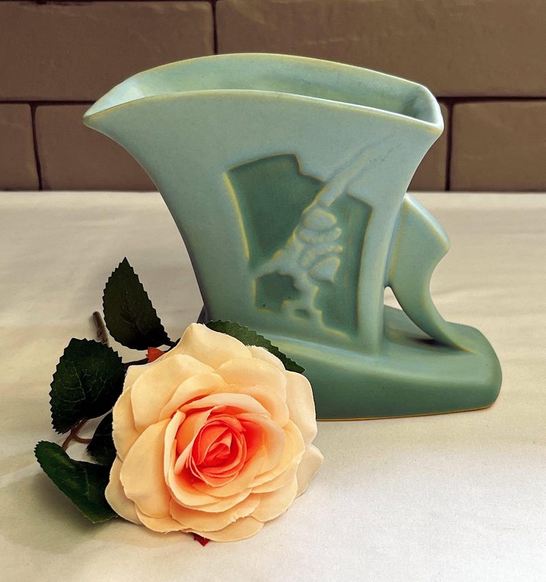 May include: A pale blue art deco vase with a relief design, next to a peach-coloured rose with green leaves. The vase has a unique shape and a matte finish, suitable for home decor.