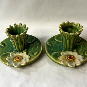 May include: Two ceramic candle holders. Each has a green lily pad base with a white and yellow flower. The candle holders are green and shaped like a flower. The candle holders are on a white surface.