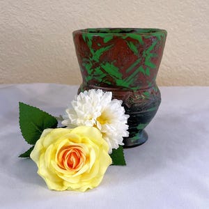May include: A handcrafted ceramic vase with a unique design. The vase features a dark brown base with green brushstrokes and a black rim. A yellow rose and white flower with green leaves are placed in front of the vase.