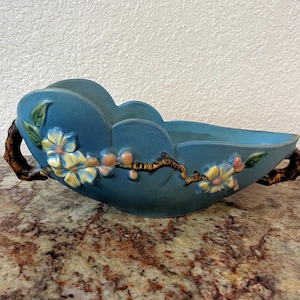 May include: A teal ceramic decorative bowl with a scalloped rim and branch-like handles. The bowl is adorned with embossed floral and branch designs in yellow, orange, and green. The bowl is approximately 12 inches long.