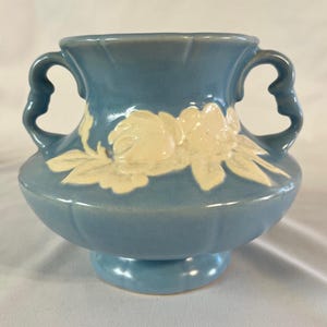 May include: A blue ceramic vase with two handles and a pedestal base. The vase features a cream-colored floral design on the front. The vase is approximately 7 inches tall and 6 inches wide.