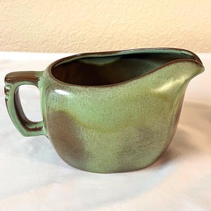 Pode incluir: Uma molheira de cerâmica com pega e bico. O exterior apresenta uma mistura de tons verdes e castanhos, com um acabamento brilhante. O interior é castanho escuro. A molheira foi concebida para servir molhos.