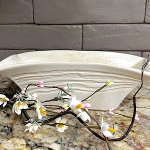 May include: A rectangular off-white ceramic planter with a flared rim and horizontal textured lines along the sides. In front of the planter lies a decorative branch featuring small white flowers with yellow centers and tiny pink buds.