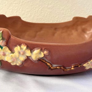 May include: A decorative, oblong, ceramic bowl in a muted rose color. The bowl features a floral design with yellow and white flowers, green leaves, and gold accents. The handles are brown and branch-like.