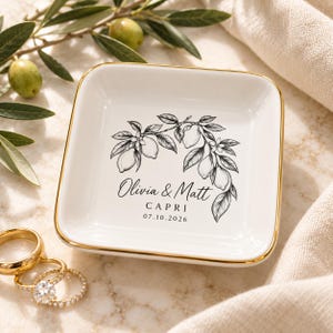 Puede incluir: Un plato cuadrado de cerámica blanca con un borde pintado en oro. El centro presenta una ilustración en tinta negra de una rama de limón con hojas y flores, junto con el texto impreso Olivia & Matt, CAPRI y la fecha 07.10.2026.